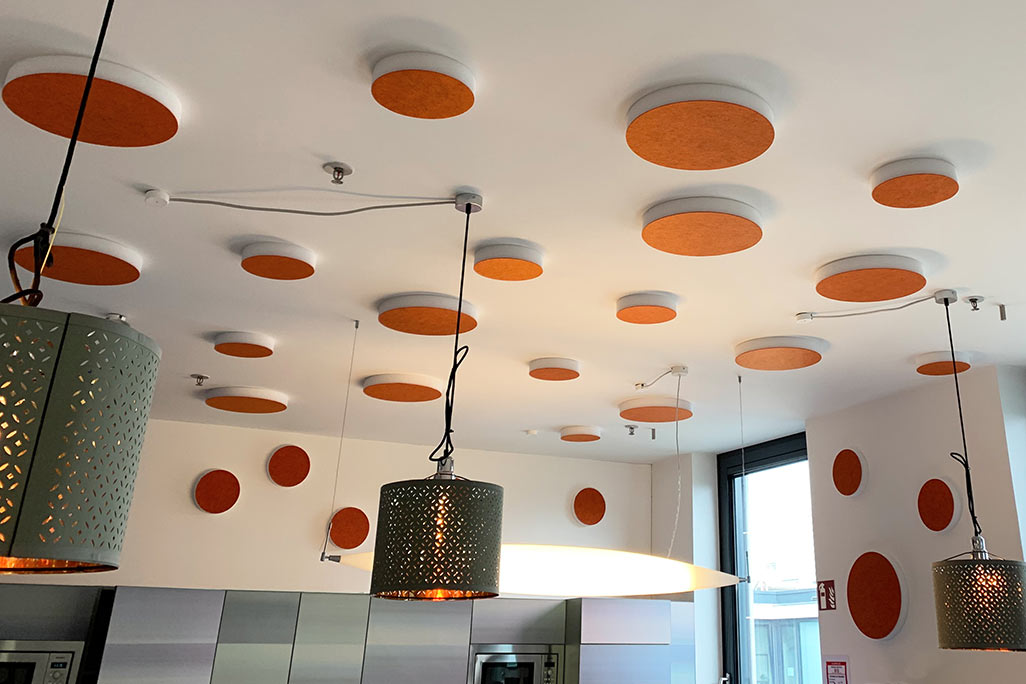 soni ELEMENT CIRCLE About 20 colored sound absorbers in the form of a large and a small circle (soni ELEMENT CIRCLE) mounted on a ceiling and a wall in the color orange in a white kitchen/breakfast room of a company.