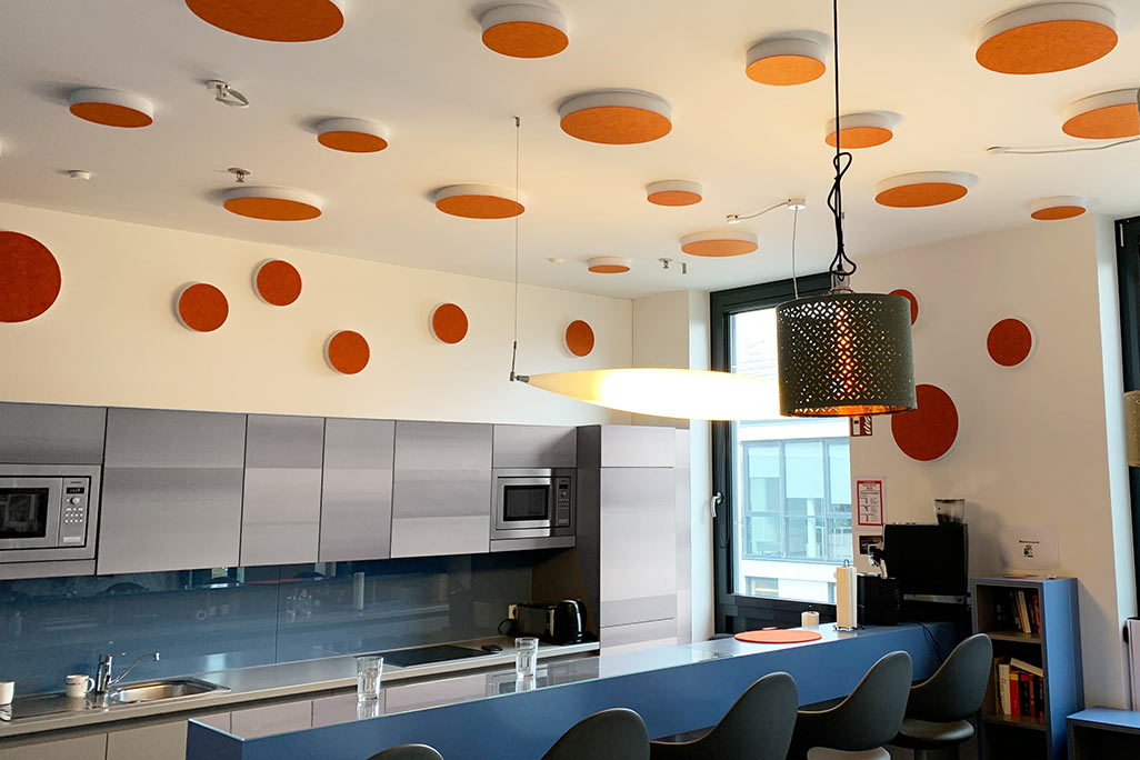 soni ELEMENT CIRCLE About 20 colored sound absorbers in the form of a large and a small circle (soni ELEMENT CIRCLE) mounted on a ceiling and a wall in the color orange in a white kitchen/breakfast room of a company.