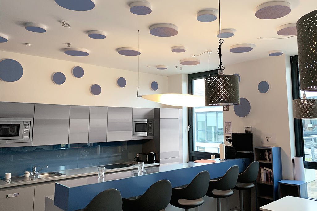 About 20 colored sound absorbers in the form of a large and a small circle (soni ELEMENT CIRCLE) mounted on a ceiling and a wall in the color blue in a blue and white kitchen/breakfast room of a company.